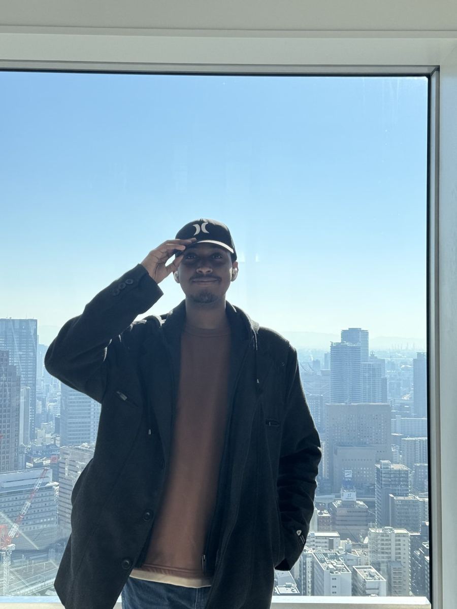 Rayan tipping his cap in front of a floor-to-ceiling window over a city skyline